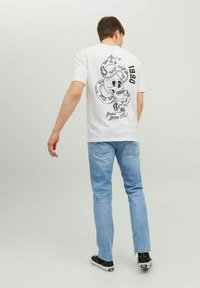 Camiseta blanca con un gráfico negro de una calavera con serpientes. Combinada con unos jeans azul claro y zapatillas negras. Diseño sencillo, estilo casual.