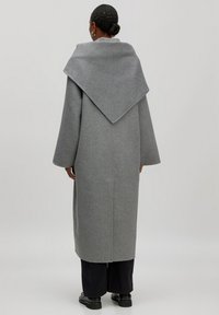 Langer grauer Wollmantel mit weitem, spitzem Kragen, abfallenden Schultern und geradem Schnitt, von hinten gezeigt. Der Stoff wirkt weich und strukturiert.