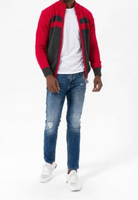 Rood en grijs gebreid jack met geribde manchetten, gecombineerd met versleten blauwe jeans en witte sneakers, met daaronder een eenvoudig wit T-shirt.