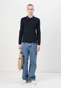 Μαύρο μακρυμάνικο πουκάμισο πόλο με αντίθετο γιακά, συνδυασμένο με ανοιχτό μπλε φαρδύ τζιν και μπεζ τσάντα. Λευκά αθλητικά παπούτσια ολοκληρώνουν την εμφάνιση.