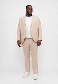 Traje de lino color beige claro con una chaqueta de un solo botón y pantalones a juego. Combinado con una camiseta blanca y zapatillas blancas.