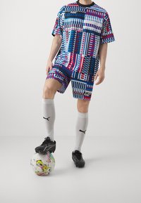 Puma urheilu-paita ja shortsit sinisissä, punaisissa ja valkoisissa geometrisissa kuvioissa, yhdistettynä valkoisiin polvisukkiin ja mustiin nappuloihin kuviollisen jalkapallon päällä.
