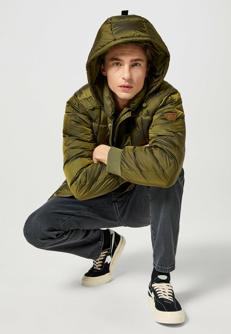 Párnázott olajzöld dzseki kapucnival, cipzárral és bordázott mandzsettákkal, sötét farmerral és fekete sportcipővel, fehér díszítéssel párosítva.