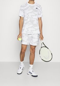 Męski tenisista ubrany w szaro-camo koszulkę i szorty, w białych butach i skarpetach Adidas, trzymający piłkę tenisową i rakietę na jednolitym tle.