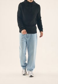 Svart stickad huvtröja med krage, i kombination med ljusblå baggy-jeans och gråa sportskor, mot en neutral bakgrund.