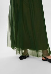 Zöld áttetsző maxi szoknya, amely légies textúrával és lebegő kialakítással rendelkezik, rétegzett anyagból készült, fekete lapos slip-on cipőkkel párosítva.