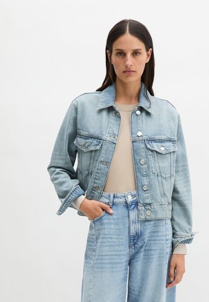 Vrouw draagt een lichtblauw spijkerjack en spijkerbroek met een beige longsleeve, staand tegen een effen witte achtergrond, met haar hand in haar zak.