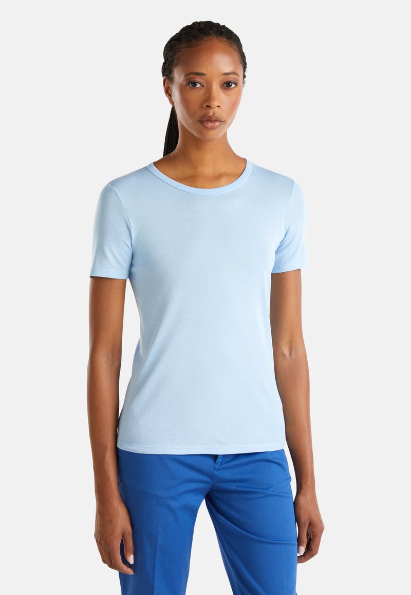 Žena s pletenými vlasy, oblečená v světle modrém tričku s krátkým rukávem a modrých kalhotách, stojící na jednobarevném bílém pozadí.