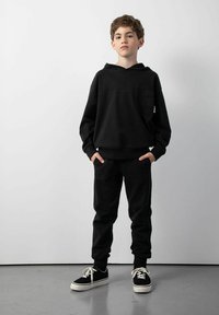 Chłopiec stojący z rękami w kieszeniach, ubrany w czarną bluzę z kapturem, czarne dresy i czarne sneakersy z białymi podeszwami i sznurowadłami na tle gładkiej ściany.