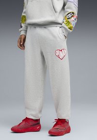 Pantalones deportivos grises de forro polar con bolsillos, que presentan un logo de corazón rojo en el muslo izquierdo. Combinados con zapatillas deportivas de color rojo brillante con parte superior texturizada.