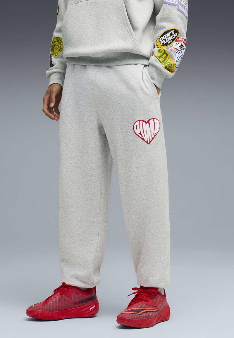 Pantalones deportivos grises de forro polar con bolsillos, que presentan un logo de corazón rojo en el muslo izquierdo. Combinados con zapatillas deportivas de color rojo brillante con parte superior texturizada.