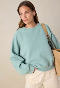 Sweatshirt oversize en vert d'eau clair avec des poignets et un ourlet côtelés, associé à un pantalon couleur crème. Le tissu semble doux et confortable.