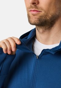 Felpa blu con zip, dotata di colletto a coste e fodera interna testurizzata, mostrata parzialmente aperta per mettere in evidenza la morbidezza del tessuto.