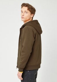Brun zip-hoodie med stor hætte, frontlomme og ribbede manchetter. Har en gul detalje på indersiden af håndleddet. Blødt, struktureret stof.