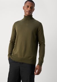 Maglione a collo alto verde oliva in tessuto a maglia liscia. Presenta maniche lunghe e un colletto e orlo a coste. Indossato con pantaloni cargo neri.