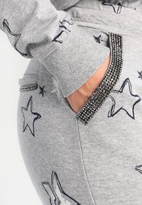 Hand in der Tasche einer grauen Jogginghose mit maritimen Sternenprints und Taschen- sowie Bunddetails mit Strassbesatz an den entsprechenden Ärmeln des Sweatshirts.