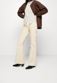 Bruine gewatteerde jas over een gestreept wit shirt, gecombineerd met beige uitlopende broek en zwarte blokhaklaarzen. Eenvoudig en modern ontwerp.