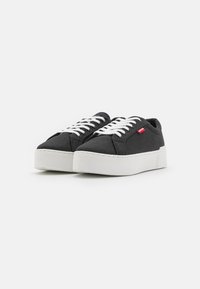 Zapatillas de lona negras con cordones blancos, punta redonda y suela de plataforma de goma blanca gruesa. Presenta un pequeño logo rojo en el lateral.