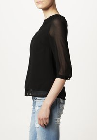 Svart, genomskinlig blus med trekvartsärmar, rund halsringning och ett strukturerat mönster vid kragen. Bärs med ljusblå, nedslitna jeans.