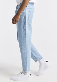 Jeans de mezclilla azul claro con un corte recto, con bolsillos delanteros y bolsillos traseros con parche. Combinados con zapatillas blancas y calcetines grises.