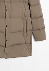 Giacca a vento marrone con un design trapuntato, dotata di tasca laterale e bottoni a pressione. Realizzata in materiale leggero e liscio.