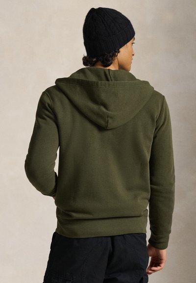 Φούτερ σε ελιά πράσινο με κουκούλα με κορδόνι, μακριά μανίκια και εφαρμοστή ραφή. Φορεμένο με μαύρο πλεκτό σκούφο. Μαλακό, υφασμάτινο με υφή.