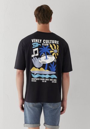 Mann trägt schwarzes T-Shirt mit bunter Vinyl Culture Club-Grafik, Palme, Sonne und Musiknoten, kombiniert mit blauen hochgekrempelten Jeansshorts.