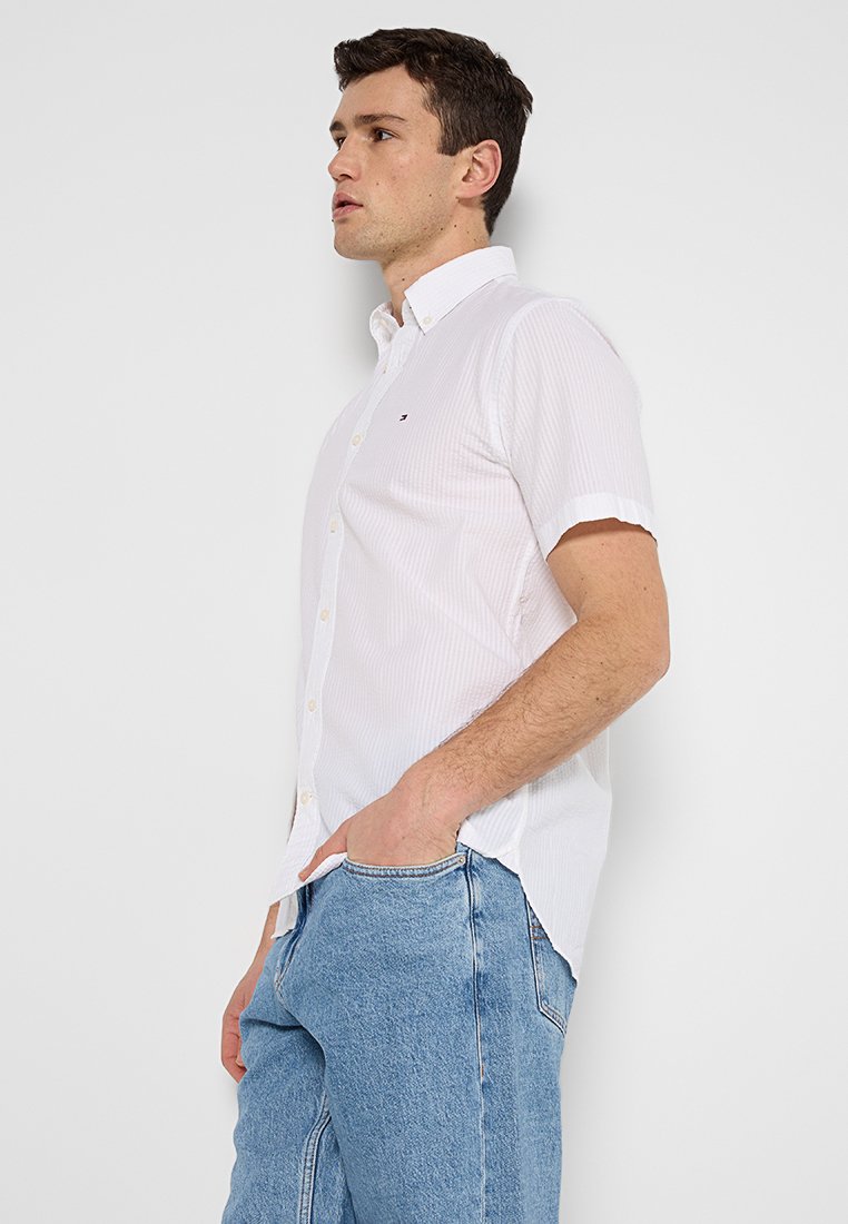 Chemise blanche à manches courtes avec un motif à rayures discret et un petit logo sur la poitrine, accompagnée d'un jean en denim bleu clair.