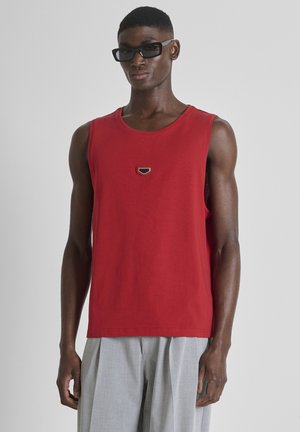 Man draagt zwarte zonnebril, rood mouwloos shirt met klein embleem en lichtgrijze geplooide broek, staand tegen een effen achtergrond.