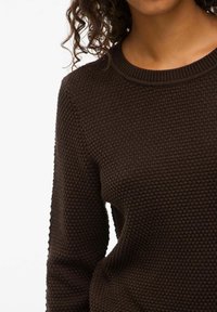 Brūns teksturēts džemperis ar apaļu kakla izgriezumu, kas raksturojas ar medus kūciņu adījumu. Audums izskatās mīksts un ir lietojams ar ievērojamu stiepšanos.