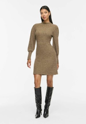 Kvinde iført en brun kabelstrikket sweaterkjole med lange puffærmer og sorte knælange støvler med hæl, stående på hvid baggrund.