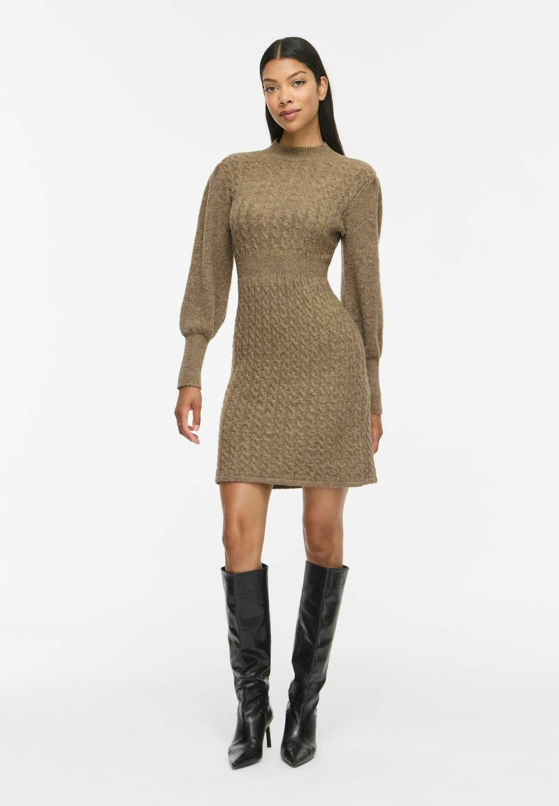 Femme portant une robe-pull marron en maille torsadée avec de longues manches bouffantes et des bottes noires à talons hautes jusqu'aux genoux, debout sur un fond blanc.