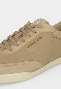 Μπεζ αθλητικό παπούτσι Michael Kors με κορδόνια μπροστά, χρυσό λογότυπο στο πλάι και λευκή σόλα, με ραφές και λεπτομέρειες σουέντ.