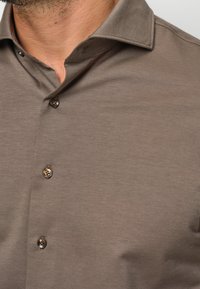 Camicia marrone con colletto a punta, realizzata in tessuto testurizzato e dotata di quattro bottoni rotondi, due dei quali sono visibili.