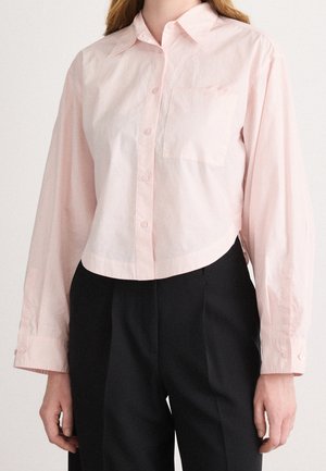 Vaaleanpunainen pitkähihaisen kauluspaita, jossa on rintatasku, puettu sisälle korkeavyötäröisiin mustiin housuihin seisovan henkilön yllä.