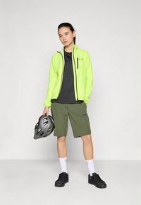 Giacca leggera giallo brillante, maglietta nera, pantaloncini verde oliva, calzini bianchi e scarpe nere; tiene in mano un casco da bici grigio.