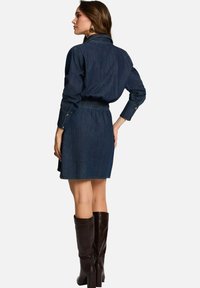 Robe en denim avec un col, taille élastique et manches longues. Couleur en denim foncé avec une jupe évasée et des poignets boutonnés. Bottes marron jusqu'aux genoux.