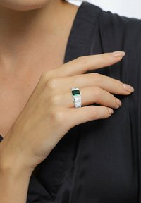Silberner Ring mit einem großen grünen rechteckigen Stein, akzentuiert durch drei kleinere klare rechteckige Steine, auf einer Hand vor einem dunklen Stoff gezeigt.
