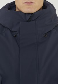 Giacca blu navy con collo alto, zip frontale e chiusure a scatto. Realizzata in tessuto liscio e opaco con accenti minimi e senza motivi visibili.