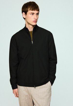 Black zip-up jacket with a stand collar, made from smooth fabric, featuring side pockets and a fitted design, worn over a green shirt.