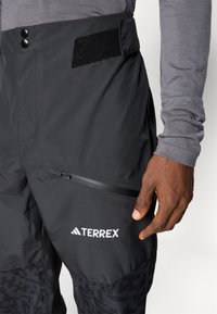 Mustat Terrex-housut, joissa on sileä pinta, vetoketjullinen tasku, tarranauha vyötäröllä ja teksturoitu alaosa, jossa on kuvioitu design.