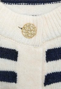 Cream knitted sweater with navy stripes, featuring a textured gold button at the neck. Soft yarn visible with ribbed collar detailing.