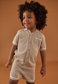 Beige gestricktes Kurzarmhemd und passende Shorts mit strukturiertem Design, ausgestattet mit einem Kragen, Knöpfen und einer Brusttasche mit gerippten Akzenten.