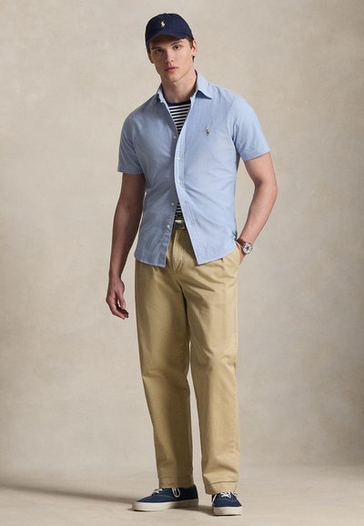 Jaunas vyras dėvi tamsiai mėlyną kepurę, šviesiai mėlyną trumpomis rankovėmis marškinius ant dryžuoto marškinėlio, smėlio spalvos kelnes, laikrodį ir mėlynas sportbačius, stovintis priešais vientisą foną.