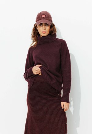Pull en maille bordeaux à col roulé associé à une jupe côtelée assortie, avec un détail de bouton sur le côté et une texture douce. Le bonnet complète la tenue.