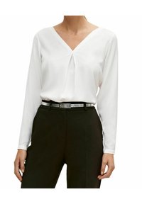Blusa blanca de manga larga con escote en V y detalle de pliegues, combinada con pantalones oscuros y un cinturón negro con acentos plateados. Textura de tela suave.