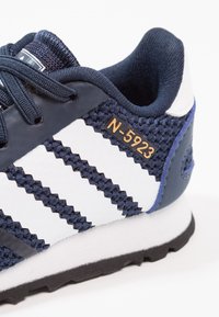 Marinblå sneakers med vitt randigt mönster, strukturerad tyg och en liten guld "N-5923"-etikett på sidan. Mjuk gummisula.