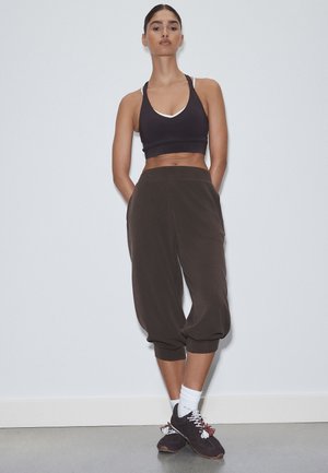 Frau trägt schwarzen Sport-BH und lockere dunkle kurze Hosen mit weißen Socken und schwarzen Sneakers, steht an einer weißen Wand.