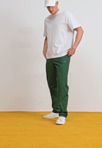 Pantaloni sportivi verzi, făcuți dintr-un material neted, asortați cu un tricou alb, pe un fond galben. Fără modele sau accente vizibile.