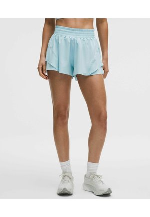 Frau trägt hellblaue, mehrlagige Sportshorts mit elastischem Bund, weiße Socken und weiße Laufschuhe, steht vor einfarbigem Hintergrund.